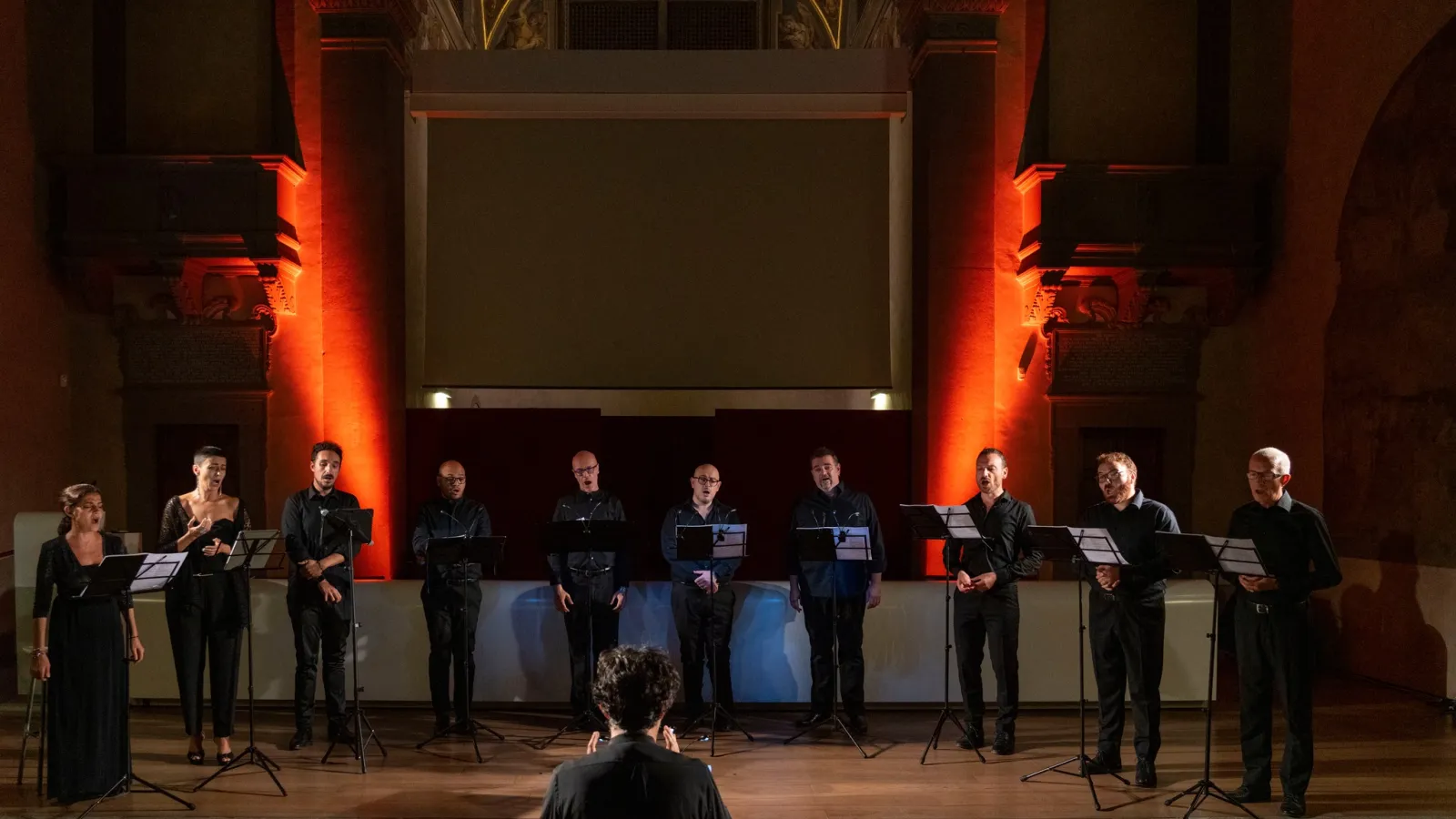 gruppo vocale composto da dieci cantanti (sette uomini e tre donne) disposti in fila su un palco, ciascuno con un leggio davanti. Davanti a loro, con le spalle alla fotocamera, si trova un direttore che sembra dirigere l’ensemble. L’ambiente è elegante, probabilmente una chiesa o una sala da concerto con architettura storica: si notano colonne, decorazioni e affreschi sul soffitto. Le pareti sono illuminate con luci calde, prevalentemente arancioni e rosse.