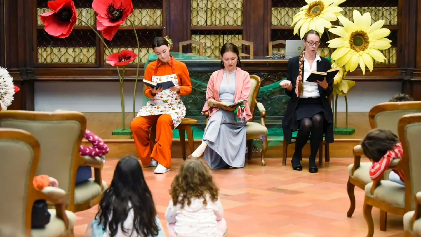 Lettura animata e laboratorio creativo per bambini con scenografia floreale (fiori giganti, papaveri rossi e margherite gialle) sullo sfondo e bambini seduti